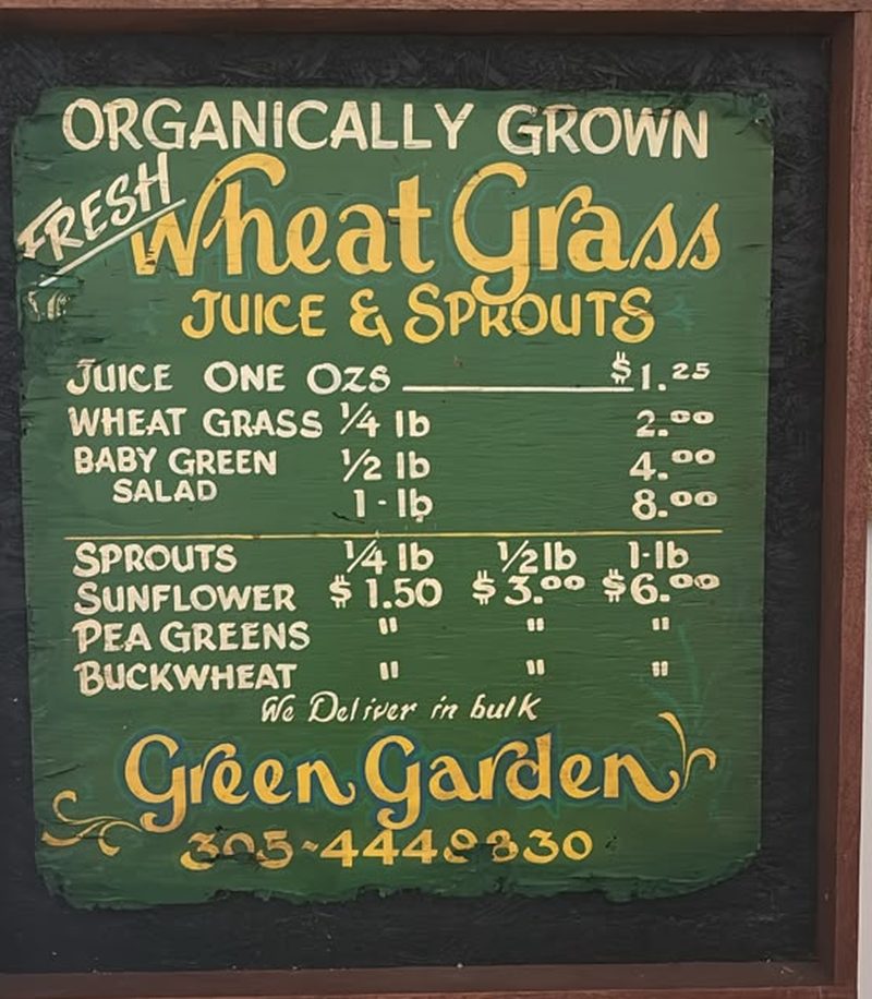 Hand-painted green price board reading Organically Grown Fresh Wheat Grass Juice and Sprouts, listing baby green salad, sunflower, pea greens and buckwheat with prices