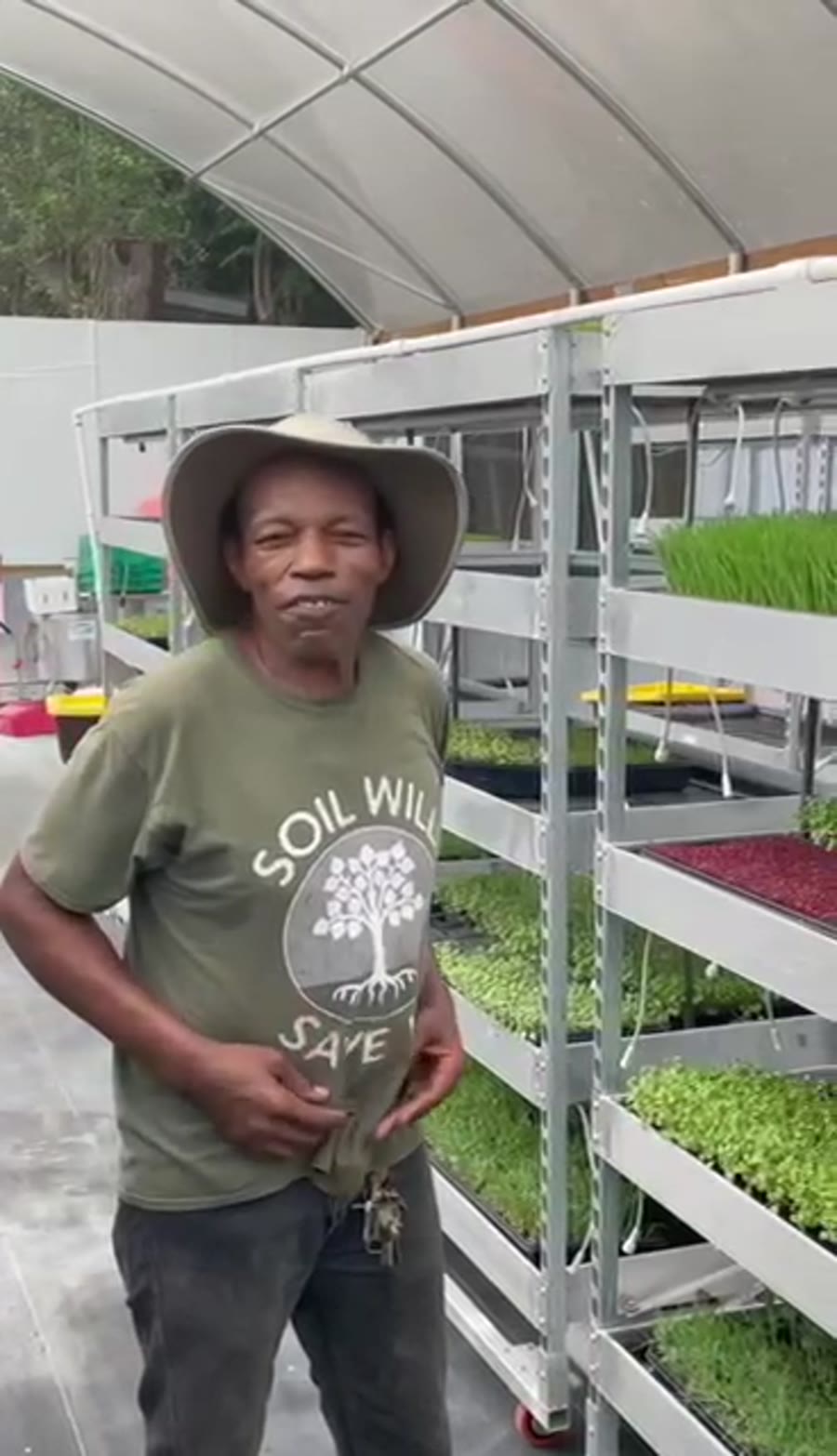 Lou Duncanson walking a greenhouse aisle beside metal racks of wheatgrass and microgreens trays